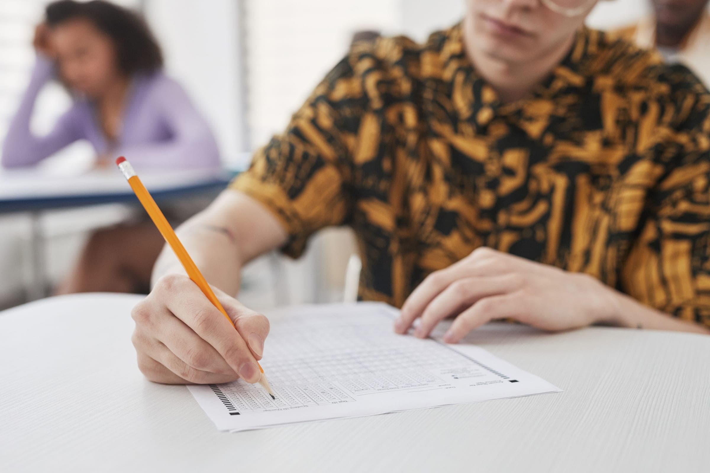 Student completing a written examination with another student working in the background.